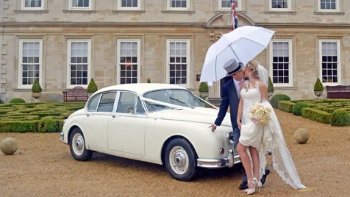 White Classic Daimler Saloon Car in front of wedding venue in Leicestershire with Bride and groom posing in front of the car kissing. Groom is holding a White Umbrella