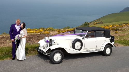 White Vintage Beauford with black roof on top of a cliff in Cornwall. Bride and Groom posing in front of the car. Blue sea as Background