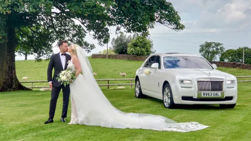 Modern white Rolls-Royce Ghost with Bride and Groom kissing next to the car . Groom wears a dark suit and Bride in White Dress with White Bridal Bouquet in her hands