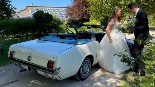 White converible american Ford mustang with Groom helping bride out of the vehicle. Groom is wearing a dark suit and the bride a White Wedding Gown.