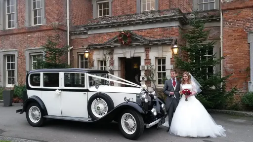 Black & Ivory Vintage Rolls-Royce Car in front of West Sussex Venue with both Bride and Groom standing in front of the car. Groom wears a Silver Suit with Burgundy tie and Bride ha