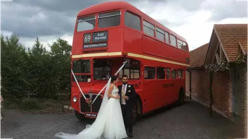Double Decker Red Routemaster bus with White Ribbons at front. Bride and Groom posing in front of the bus