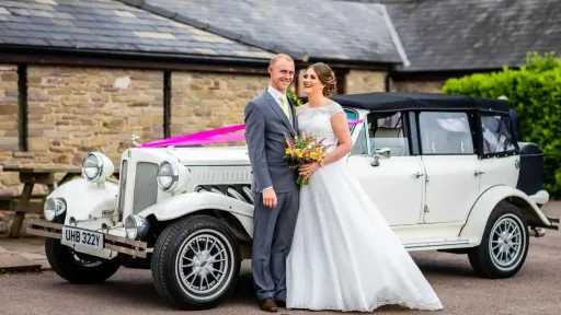 White vintage Beauford with whitewall tires, pink ribbon decoration on bonnet  and black roof outside wedding venue in Pembrokeshire. Bride and groom standing next to the car with