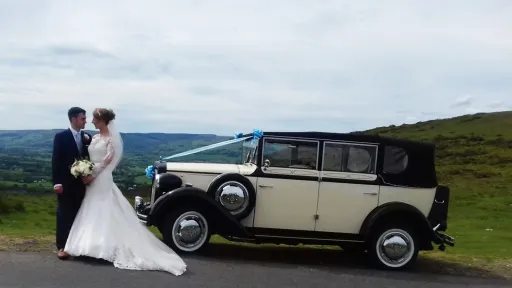 1930's Vintage Style Regent with a black Convertible roof and decorated with blue wedding ribbons on top of a cliff. Bride and Groom are standing in front of the car having their p