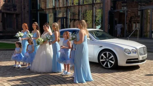 White Bentley Flying Spur with Black and Silver Alloy Wheels behingd bride wearing a white wedding dress in the middle of her bridemaids and flower girls all wearing light blue mat