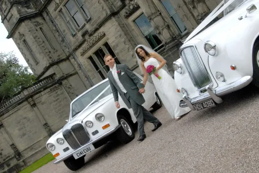 Two Classic Limousine in Northamptonshire with bride and groom standing in the middle of the cars. Bride wears a White Dress and hold Pink Flowers in her hands