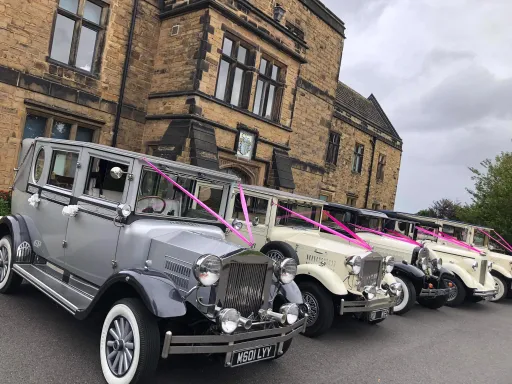 Selection of vintage cars in cream and silver in front of wedding venue in Derbyshire. All vehicles are decorated with the traditional V-Shape Ribbons on front bonnet in Pink Colour