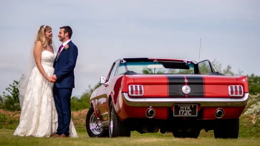 Rear view of a Convertible American Mustang with Black stripe. Bride and Groom standing by the car holding hands. Bride wears a white dress and groom has a Navy Blue Suit