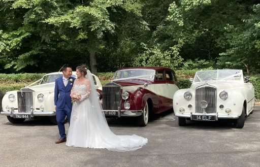 Bride and Groom posing in front of three classic cars they've hired for their Surrey wedding.  Each cars are dressed with White Ribbons