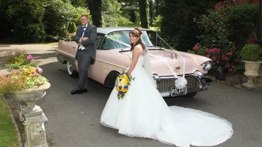 Pink American Wedding Car with white ribbon and large white bow in front of its chrome grill. Groom wears a light grey suit leaning on the vehicle while the bride wearing a long wh