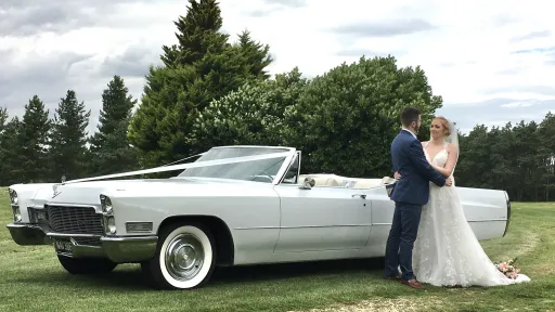 Classic White American Cadillac decorated with White Ribbons and white wall tires in a park in Suffolk with Bride and Groom standing by the car