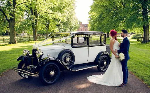 White vintage Car in Buckinghamshire in an alley with trees on both side and bride and groom standing by the vehicle. Bride wears a white dress and hold a White Bouquet of Flowers