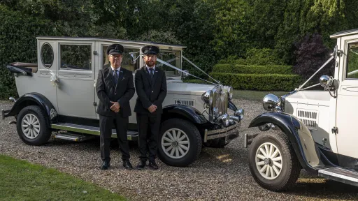 Two matching Vintage Cars decorated with White Ribbons and their chauffeurs standing by the cars with their full dark grey uniform and chauffeur's hat