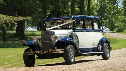 Blue & White Vintage Bramwith Limousine decorated with white ribbons driving toward Essex Wedding venue. Chauffeur wearing chauffeur's hat at the wheels and Bride and Groom at the