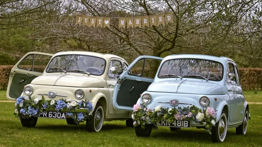Two Classic Italian Fiat 500 in Ivory and Pale Blue decorated with garland of flowers on their chrome front bumper. Both cars have white wall tires and passenger's door is open