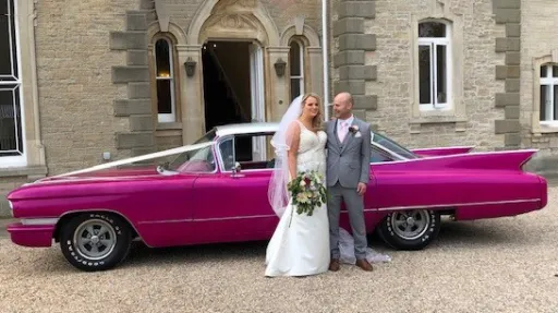 Pink Cadillac decorated with White ribbons with bride and groom standing in front of the vehicle