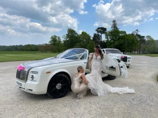 Two Rolls-Royces at venue in Somerset. Bride exiting the convertible white rolls-royce. the second vehicle just behing is a hard top rolls-royce phantom in white. Both vehicles are dressed with pink ribbons and bows