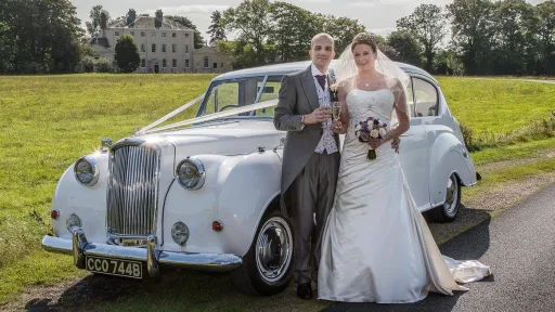 Classic Austin Princess Limousine dressed with white ribbons.. Both bride and groom standing in front of the vehicle and their Staffordshire wedding venue in background