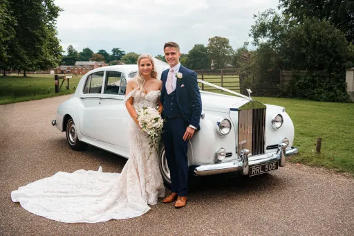 Bride and Groom posing for photos in front of the classic rolls-rocye silver cloud they've hired for their Cambridgeshire wedding. Car is dressed with traditional V-Shape white rib