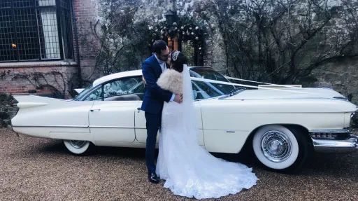 White Classic American Cadillac with white ribbons. Both Bride and Groom are kissing in front of the car