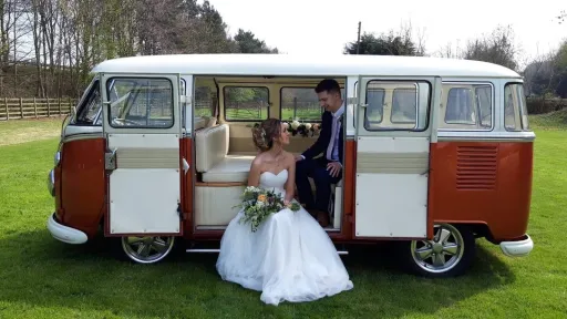 Side view of Classic VW campervan in White and Brown with its double door open. Bride and groom are sat inside the vehicle look at each others. Bride holds her bridal bouquet in he