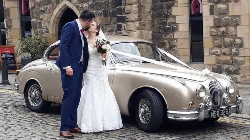 Champagne coloured Classic Jaguar Mk2 in Cumbria decorated with white ribbons. Bride and groom in front of the car kissing
