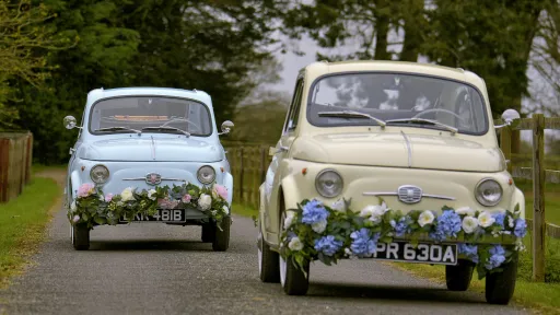 Two Classic Fiat 500 decorated with Wedding flowers on bumper following each other on Lancashire road.  Front car on the right is ivory and the rear classic Fiat on the left is in