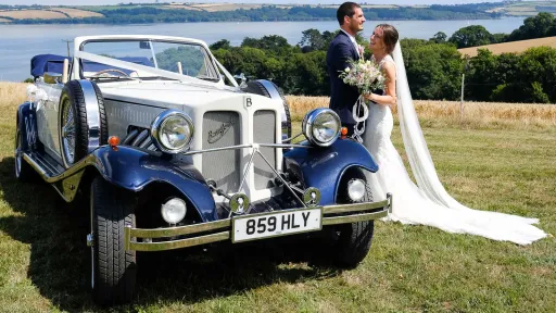 White and Blue Beauford Convertible decorated with white ribbons on the top of a cliff in Kent coastline. Bride and Groom posing next to the vehicle. Bride is holding her wedding b