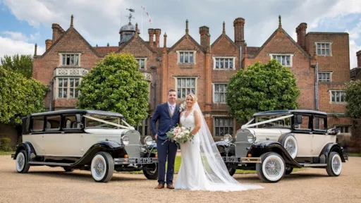 Two 1930's vintage Wedding cars decorated with white ribbons in front of Berkshire Wedding venue. Bride and Groom in the midle of both vehicles posing for their wedding photographe