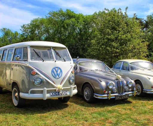 3 Classic Vehicles dressed with matching white ribbons in a park in Rutland. Left is a campervan, middle car is a Jaguar and the car on the right side is a daimler 250