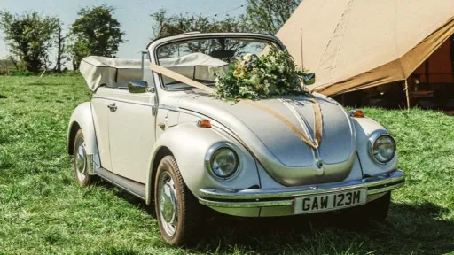 White VW beetle in a field in Warwickshire in front of the wedding marquee decorated with gold ribbons and weddiong flowers on its bonnet