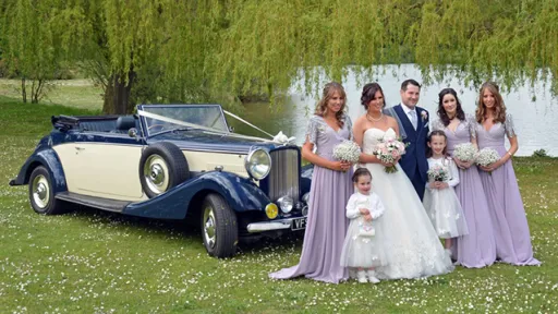 Blue & Ivory Vintage convertible car with white ribbons standing behind the Bride, Groom, Bridesmaids and their flower girls in a park under a tree in Nottinghamshire