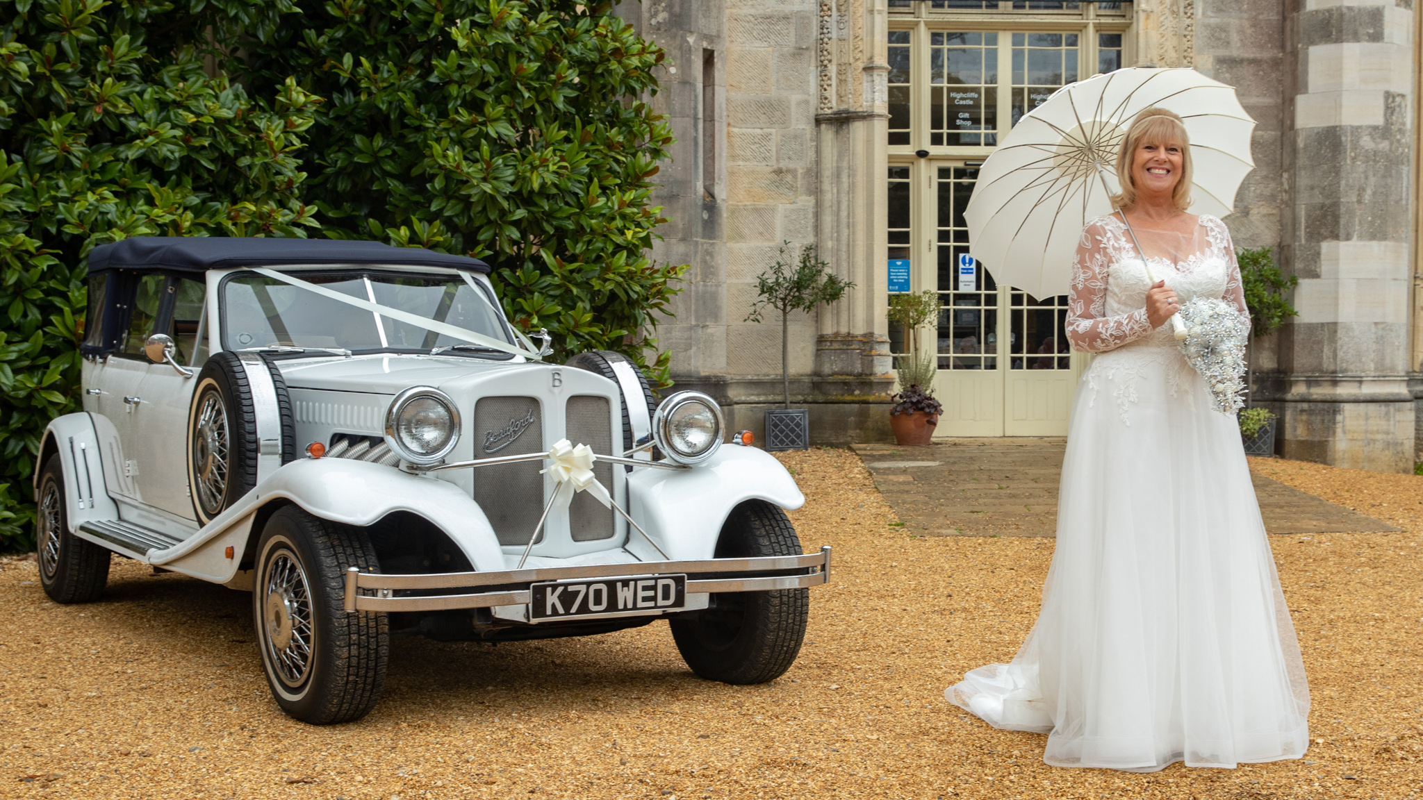 White vintage Beauford convertible with roof up decorated with white ribbon and bow at a local Faversham wedding. Smiling bride standing next to the vehicle with a white umbrella h