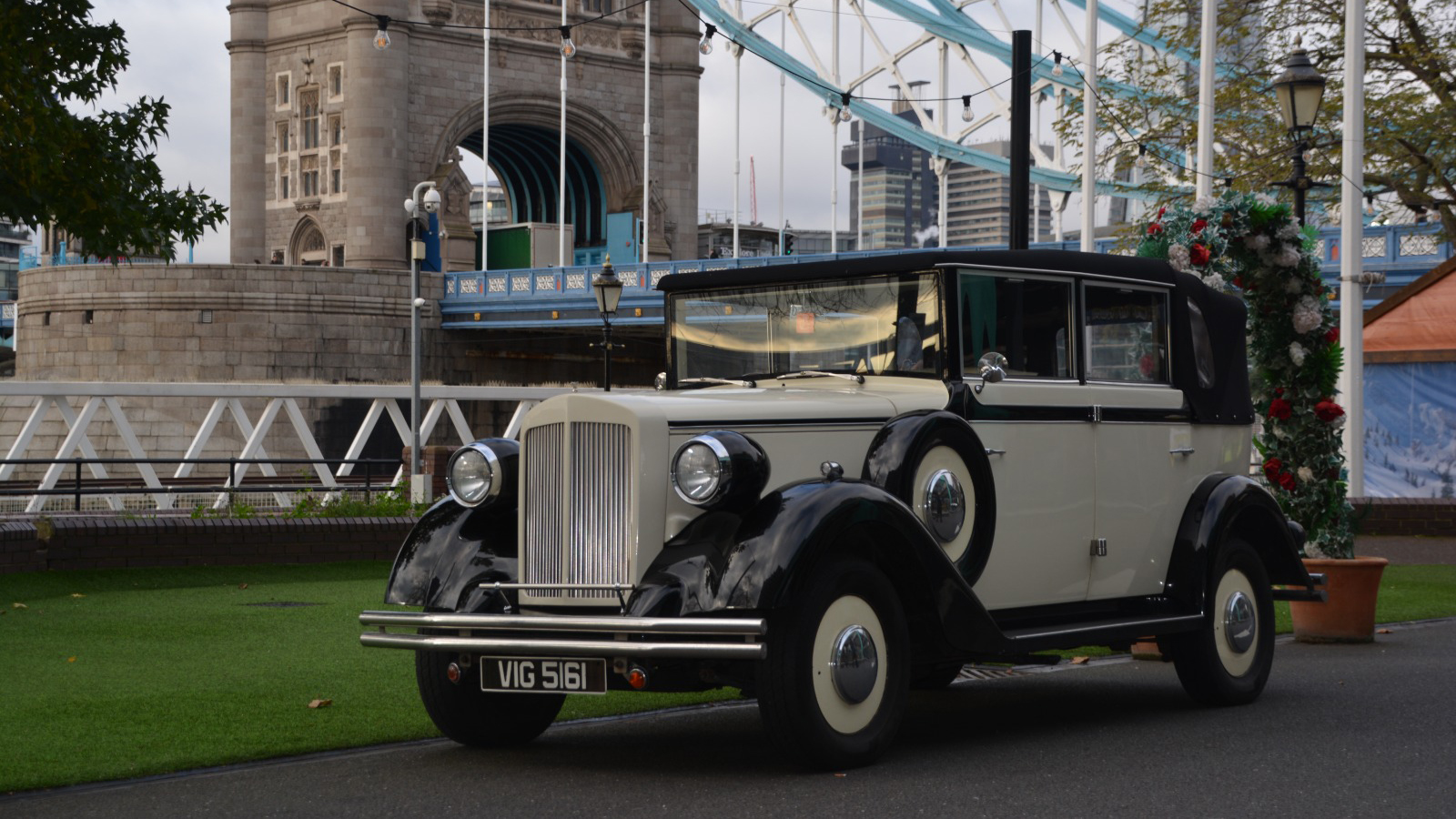 1930's vintage style regent parked in front of the London bridge. Vehicle is ivory with black wheel arches and a convertible soft-top roof.