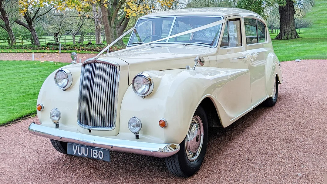 Ivory classic 7-seater Austin Princess Limousine dressed with a traditional V-shape ivory ribbons across its front bonnet. Vehicle is standing in the middle of a path in a Basildon