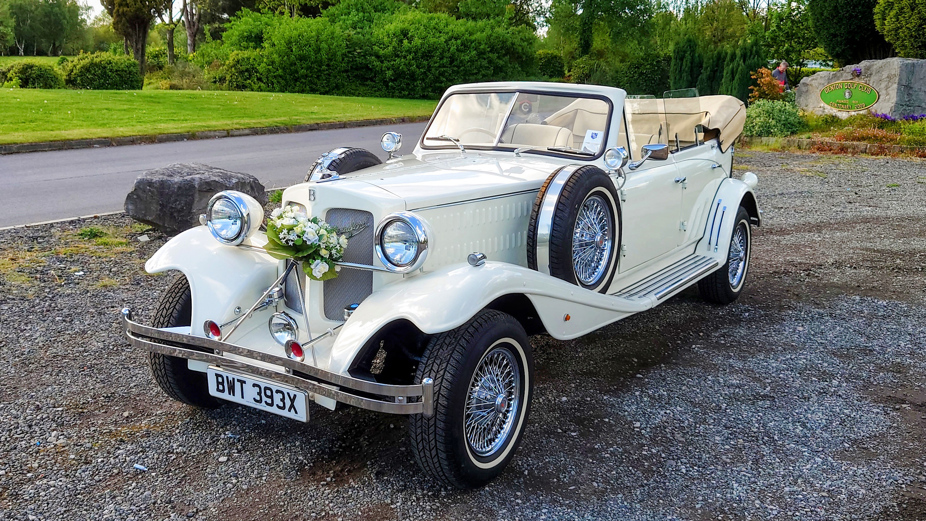 Ivory classic convertible Beauford with roof down dressed with wedding flower arrangment on its front grill. Vehicle is parked in the middle of a path in a Crewe Park.