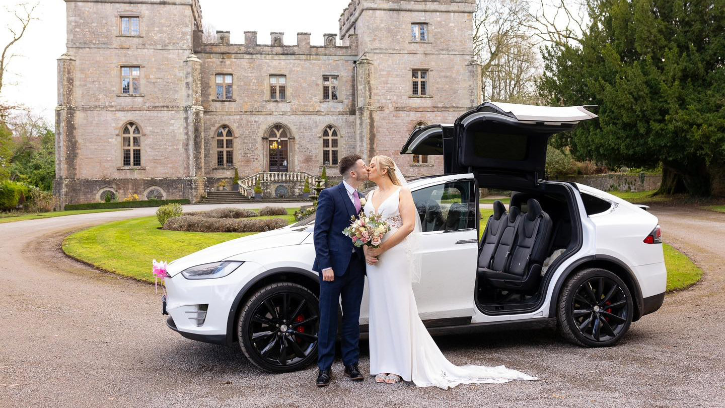 A Modern 6-seater Tesla X in White parked in front of a popular wedding venue in Newquay with its gull-wings open. Bride and Groom are standing in front of the vehicle kissing.