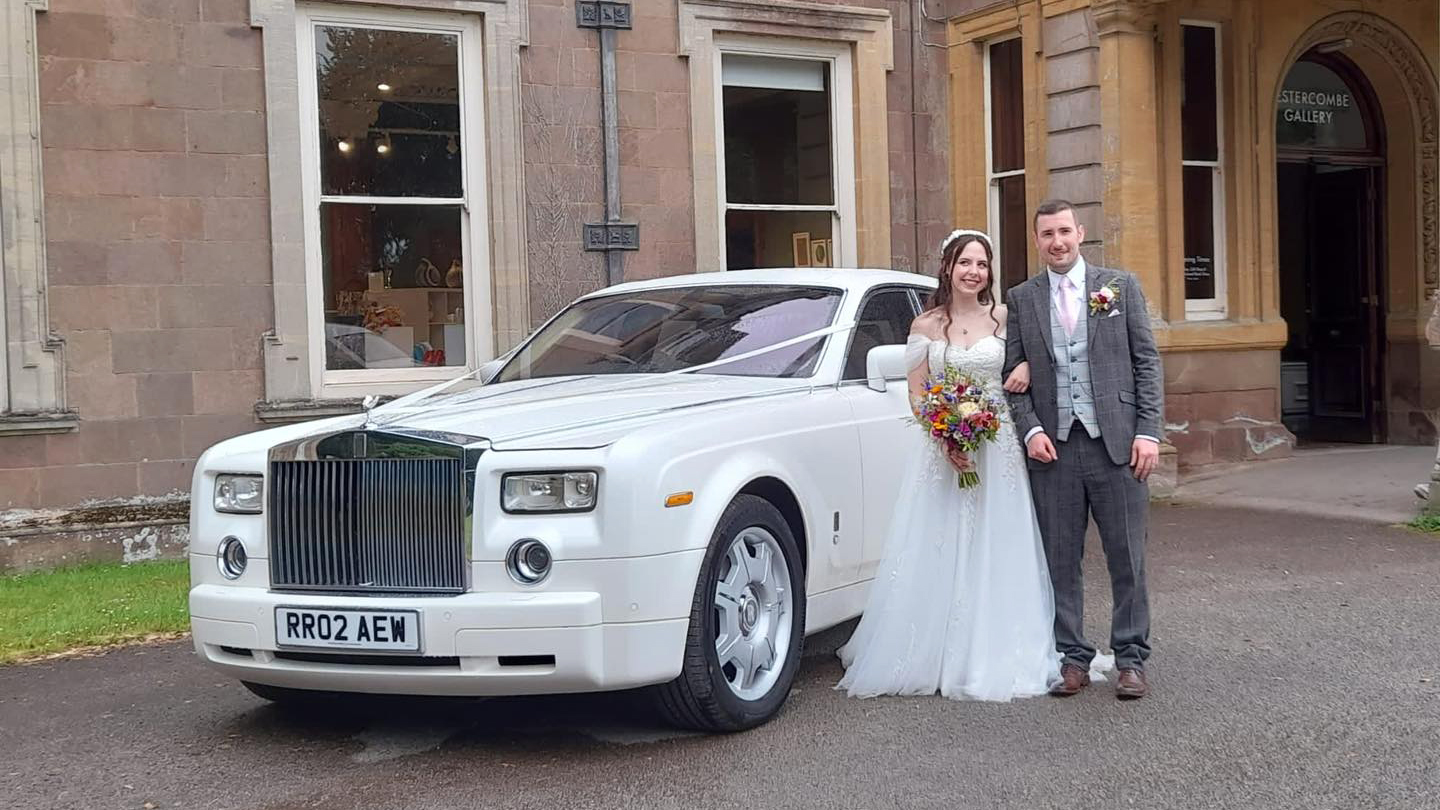 Modern white Rolls-Royce Phantom with its large chrome gril decorated with flower arrangment on top. Bride and Groom are standing by the vehicle holding each others