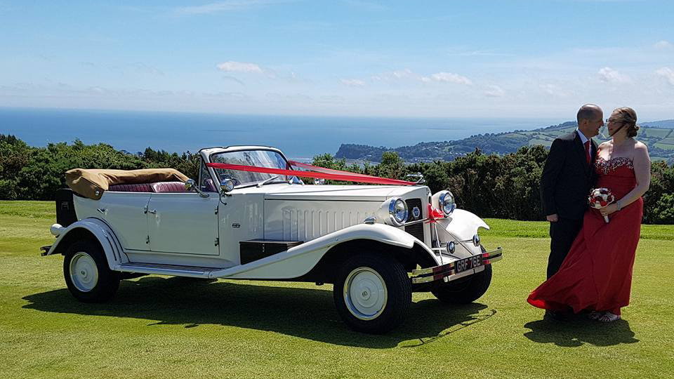 Ivory Vintage Beauford Convertible with roof down decorated with a burgundy ribbon. Bride and Groom are standing in front of the vehicle holding each others.
