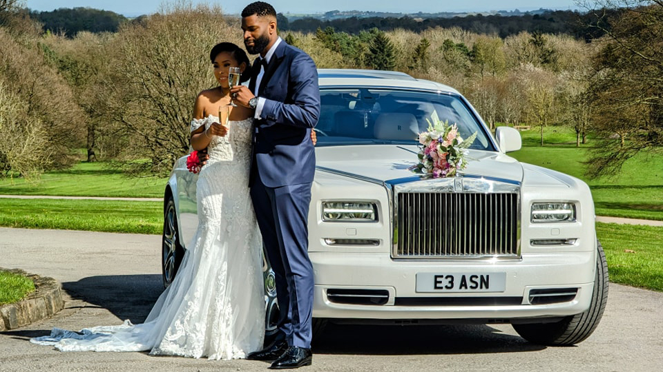 Modern white Rolls-Royce Phantom with its large chrome gril decorated with flower arrangment on top. Bride and Groom are standing by the vehicle holding each others