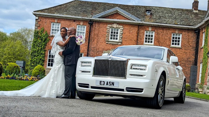 A Modern Rolls-Royce Phantom in White with Bride and Groom holding each others in front of the car posing for their wedding photographer. A popular wedding venue in Wigan can be seen in the background.