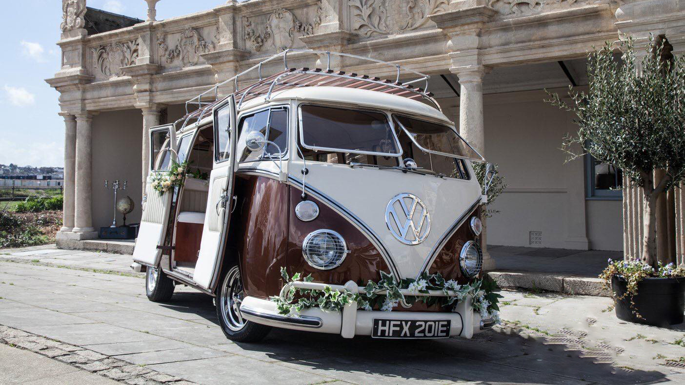 Retro Classic Volkswagen Campervan Splitscreen in Bronze and White decorated with floral arrangment on its front bumper and doors. The Camper is parked in front of a local Tiverton wedding venue.