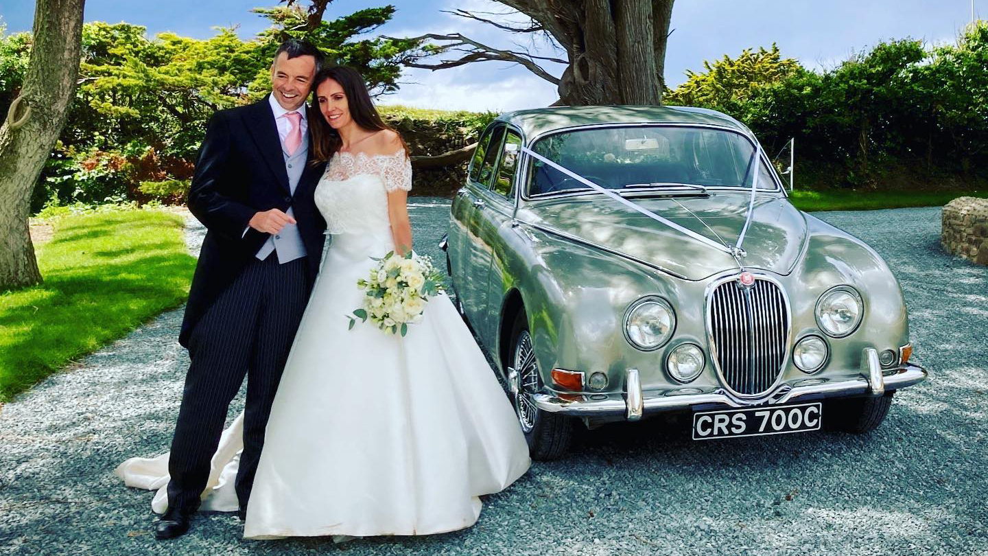 Classic Jaguar MK2 with Bride and Groom standing in front of the vehicle smiling. The Bride is holding her bridal bouquet in her left hand.  Vehicle is parked in the garden of a lo