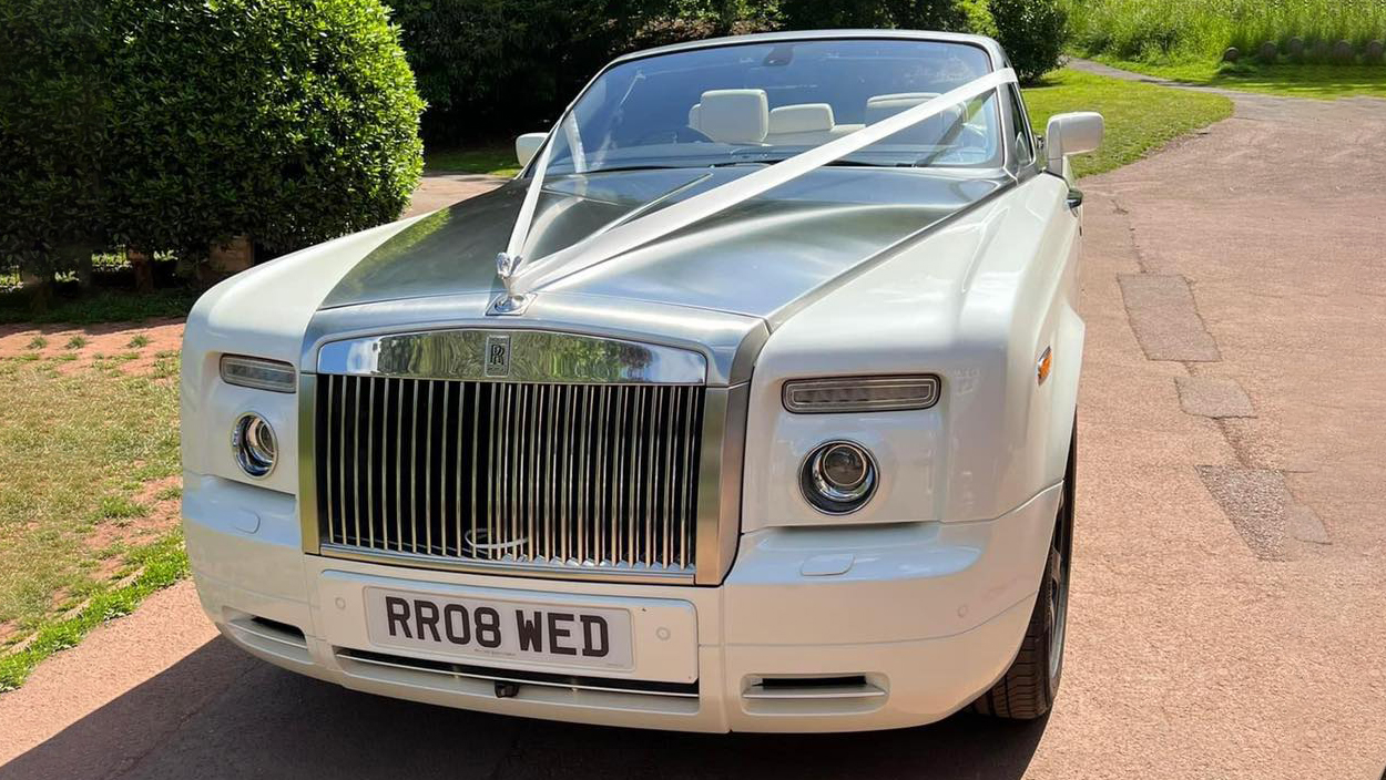 Modern white Convertible Rolls-Royce Phantom with its large chrome gril decorated with white wedding ribbons across its bonnet. The roof is down showing a cream leather interior.