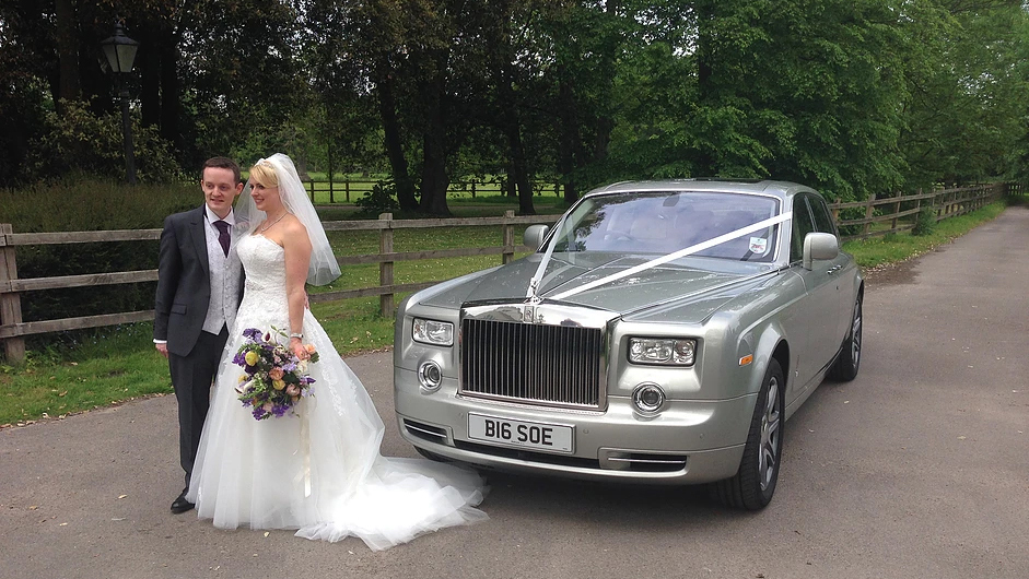 Modern Silver Rolls-Royce Phantom Series 2 decorated with traditional white ribbons. Bride and Groom are standing in front of the vehicle