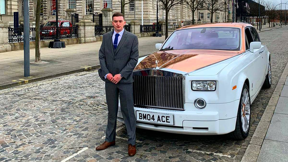 Modern white Rolls-Royce Phantom with rose gold bonnet in the street of Blackpool with its fully-suited chauffeur standing at the front of the vehicle.