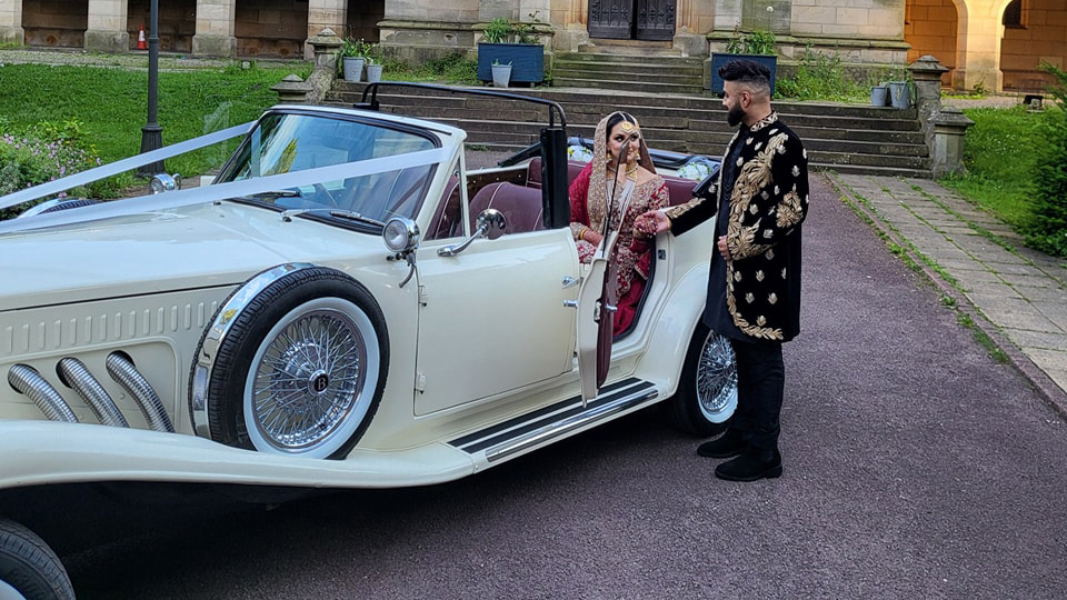 A Vintage Beauford Convertible in Ivory with Asian Bride seating in the back seat and groom standing by the side of the vehicle holding his bride's hand.