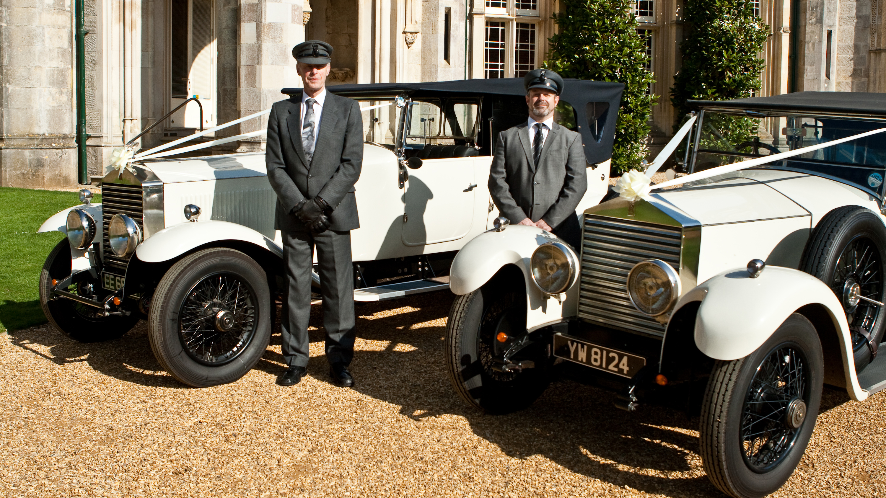 Two vintage wedding cars decorated with white ribbons and bows at a local Abbotsbury wedding. Both vehicles are parked side-by-side with their chauffeurs next to them.