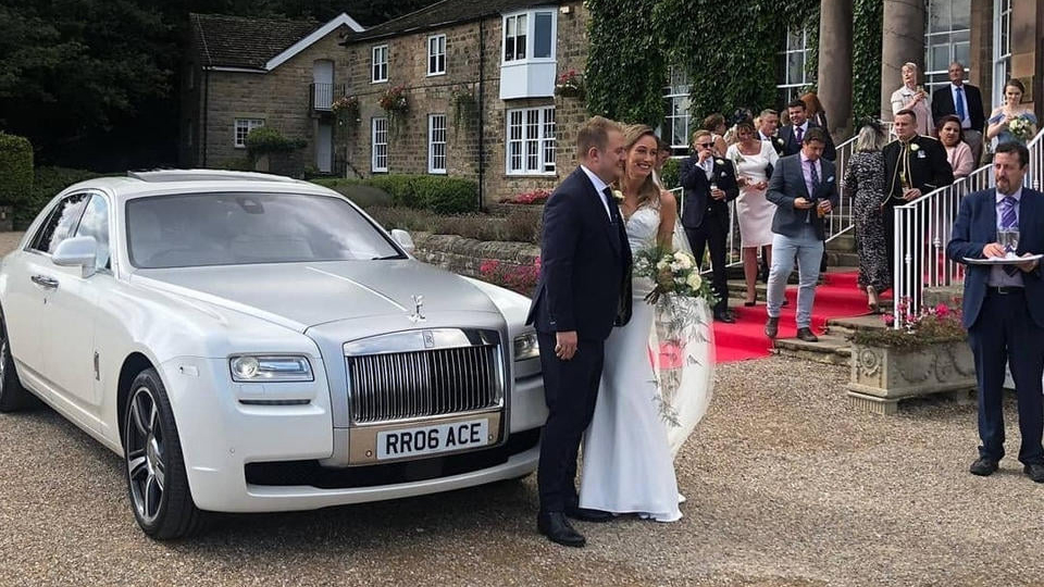 Modern White Rolls-Royce Ghost with silver bonnet. Bride and Groom are standing in front of the vehicle with their wedding guests in the background at a local Leyland wedding venue.