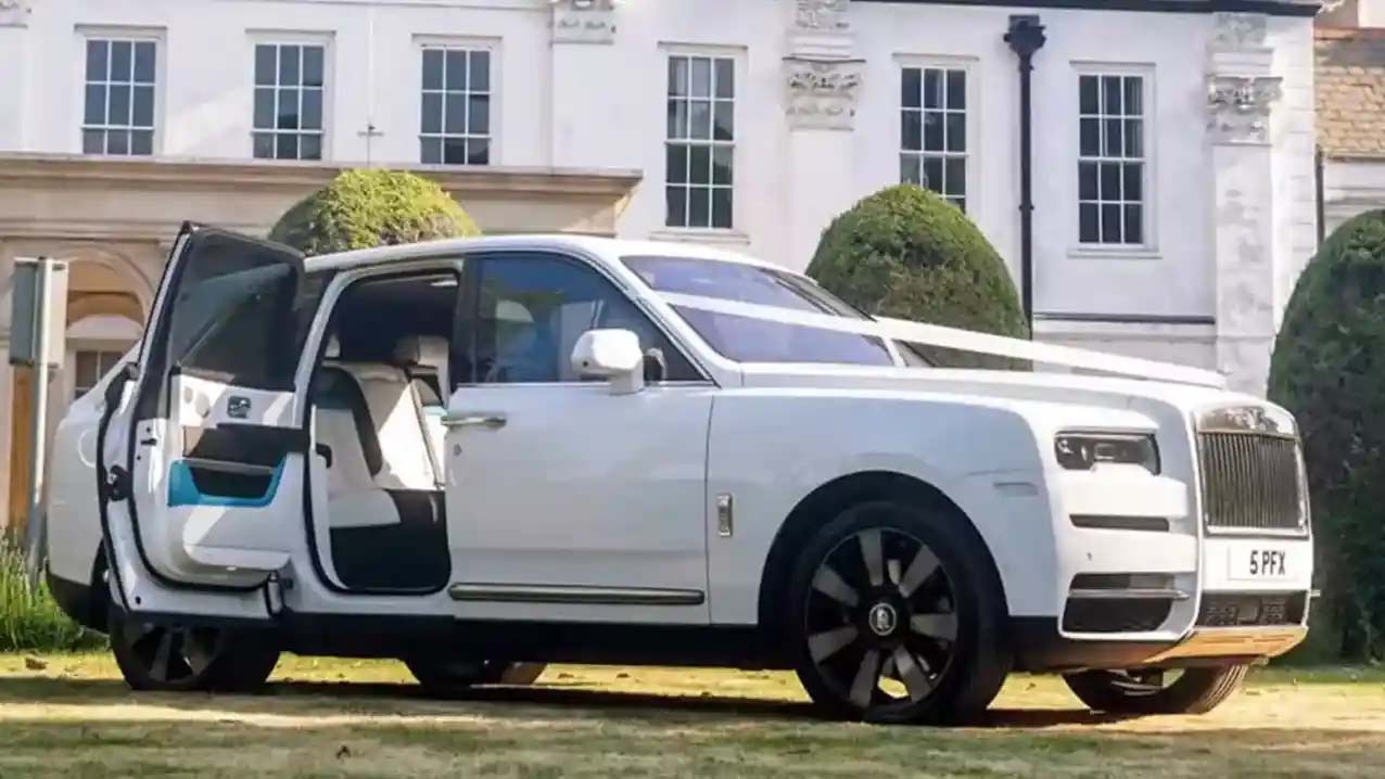 A modern white Rolls-Royce Cullinan with rear door open showing the cream leather interior in St Albans. Wedding venue can be seen in the background